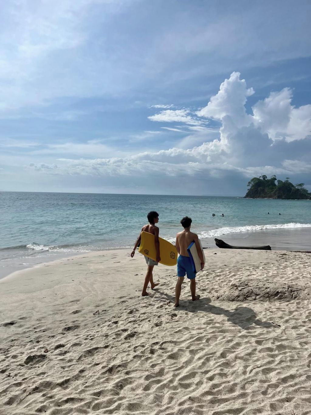 Zwei Personen laufen mit Surfbrettern über einen sandigen Strand, das Meer ist ruhig und in der Ferne ist eine Insel zu sehen.