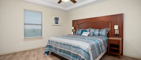 Une chambre à coucher avec un lit, un ventilateur de plafond, une table de chevet, une lampe, une fenêtre avec des stores et un tableau au mur.