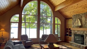 A cozy living room with large arched windows, comfortable seating, a fireplace, and a stone accent wall. - Chandos Lake Sanctuary (North Kawartha)