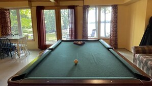 Una mesa de billar con la bola blanca en el centro, rodeada de ventanas con cortinas, una mesa de comedor con sillas y un sofá.
