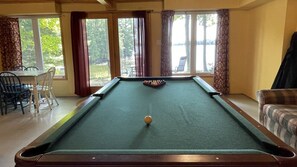 A pool table with a cue ball in the center, surrounded by windows with curtains, a dining table with chairs, and a sofa. - Chandos Lake Sanctuary (North Kawartha)
