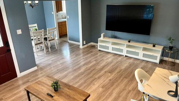 Moderni olohuone, jossa on puulattia, seinälle asennettu televisio, valkoinen mediakeskus ja ruokailutila, jossa on pöytä ja tuolit.