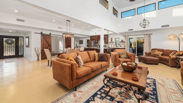 A spacious living room with a large brown sofa, a coffee table, and a patterned rug. There is a kitchen area in the background with modern appliances and a dining area with a table and chairs.