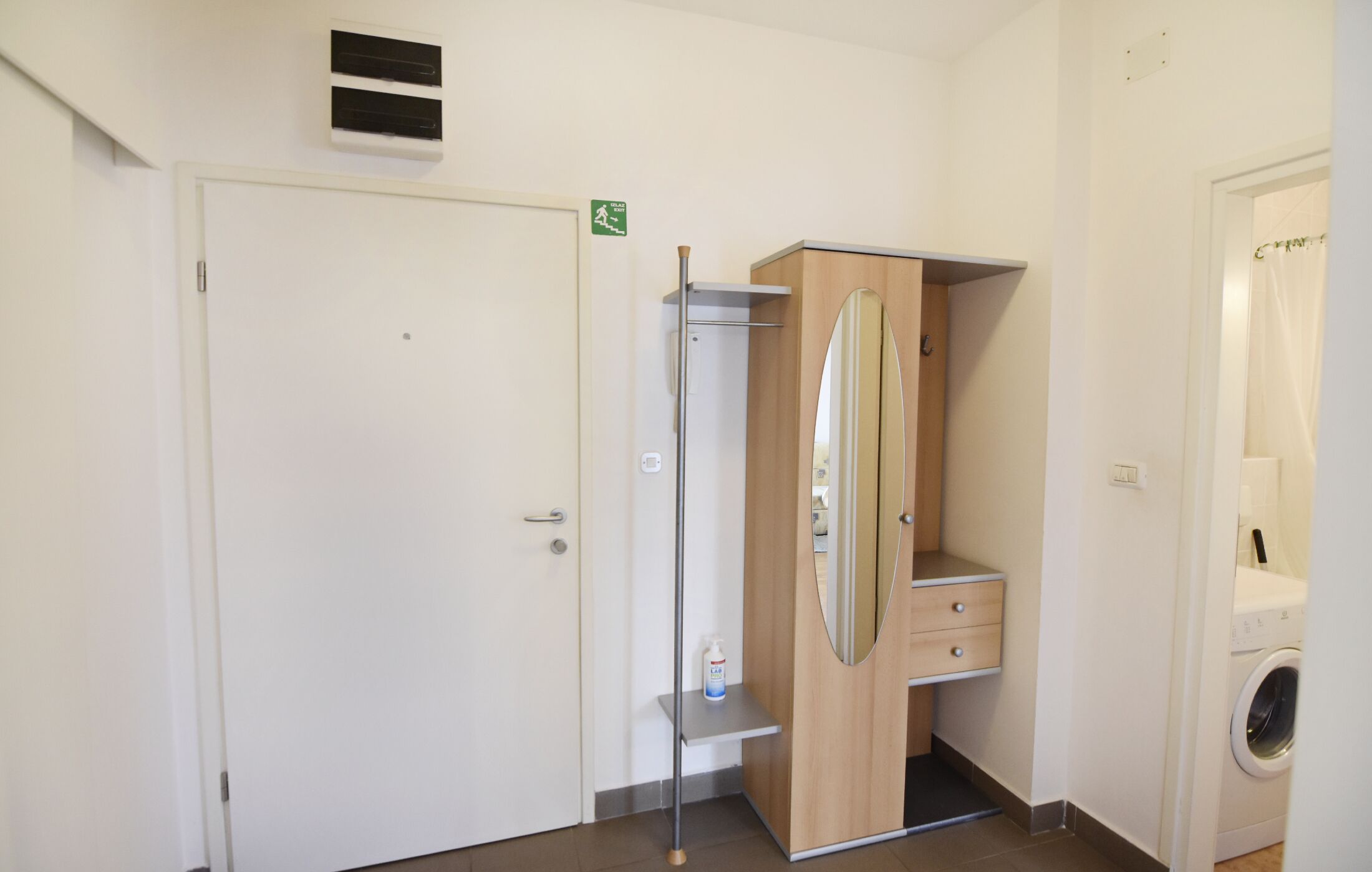 Een hotelkamer met een witte deur, een houten garderobe, een klein plankje met een fles en een wasmachine.
