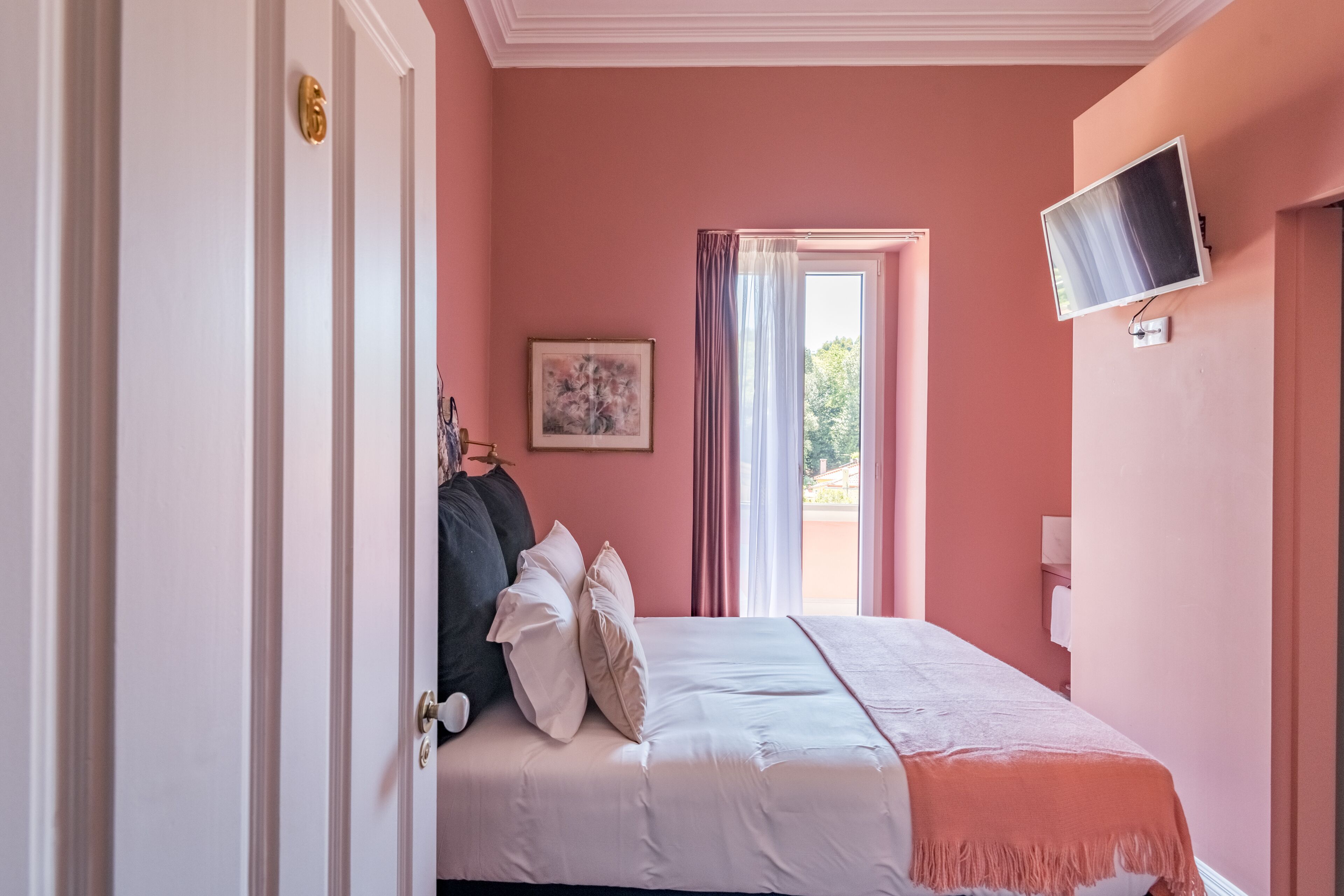 Un dormitorio con cama, televisor, un cuadro y ventana con cortinas.