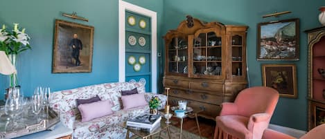 Ein Wohnzimmer mit türkisfarbenen Wänden, einem blumigen Sofa, einem rosa Sessel, einem Holzschrank und einem Glastisch.