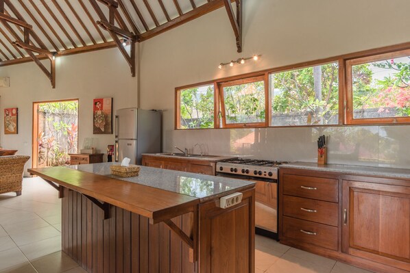 A kitchen with wooden cabinets, a central island, a gas stove, and large windows with a view of greenery. - Villa Lotus Kayuputih (Sukasada)