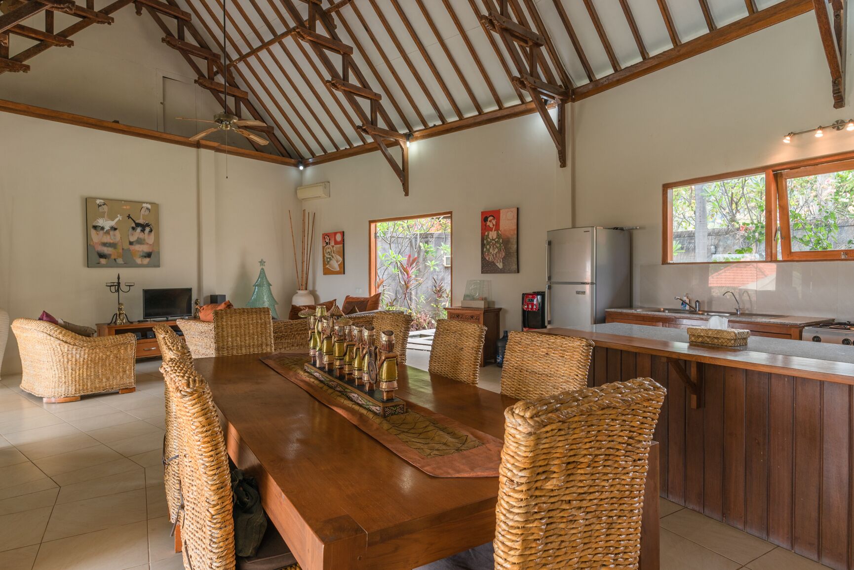 A spacious dining area with a long wooden table and wicker chairs, a kitchen with a refrigerator and sink, and a ceiling with exposed wooden beams.