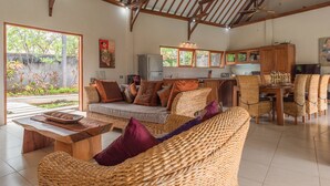A spacious living area with a sofa, dining table, and chairs, a kitchen with wooden cabinets, and a large window offering a view of greenery. - Villa Lotus Kayuputih (Sukasada)
