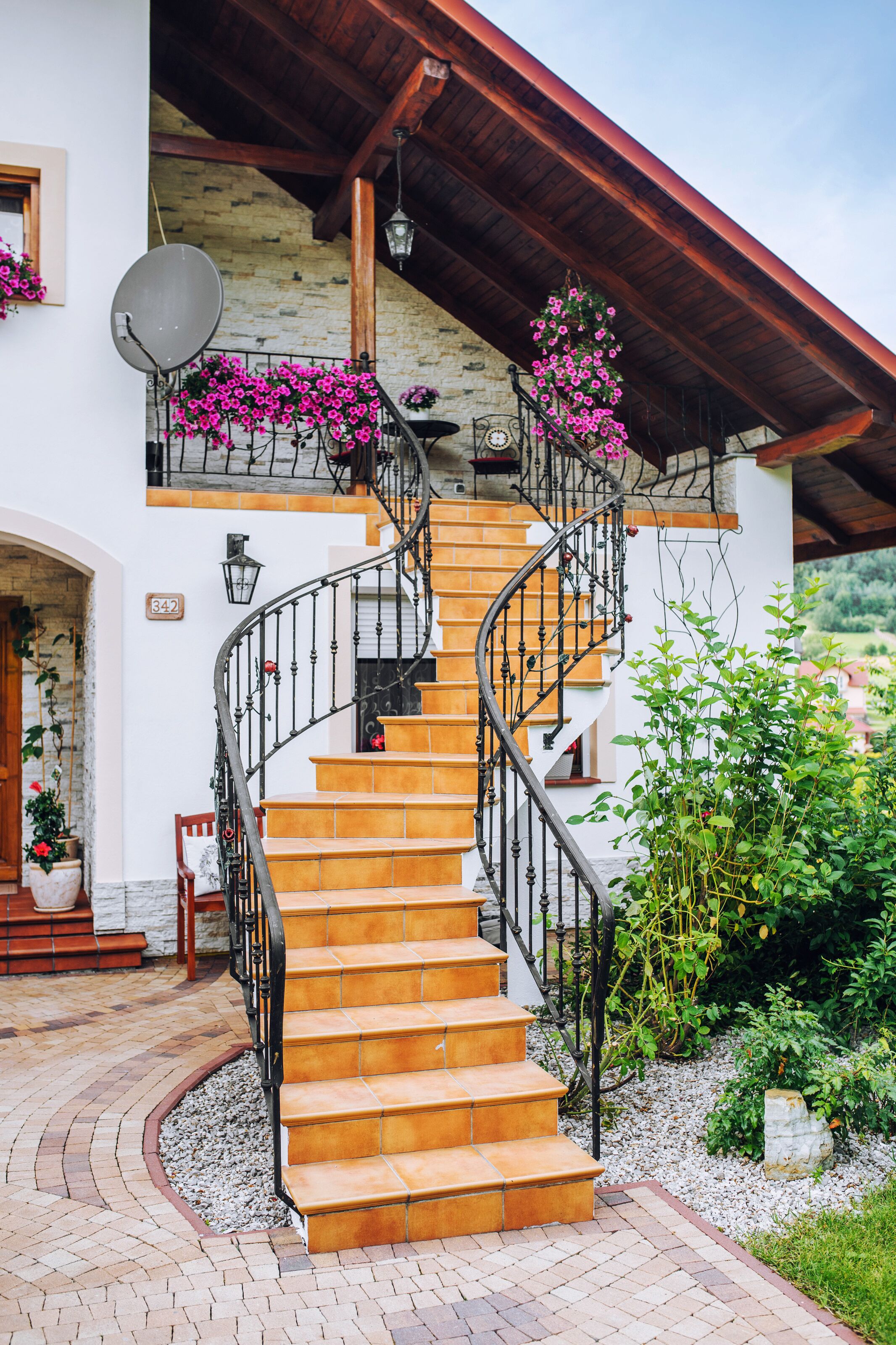 Una escalera en espiral conduce a un balcón del segundo piso con flores rosadas, una pared de piedra y una antena parabólica.