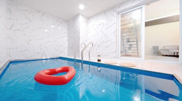 En swimmingpool med en rød redningskrans, metalstiger og en glasdør ind til et opholdsområde med en sofa og et lille bord.