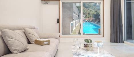 A modern living room with a sofa, a coffee table, and a large window overlooking a swimming pool.