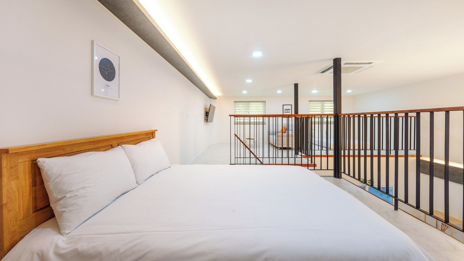 Une chambre avec une tête de lit en bois, un lit recouvert de linge de lit blanc, un tableau encadré au mur et un escalier avec une rampe noire.