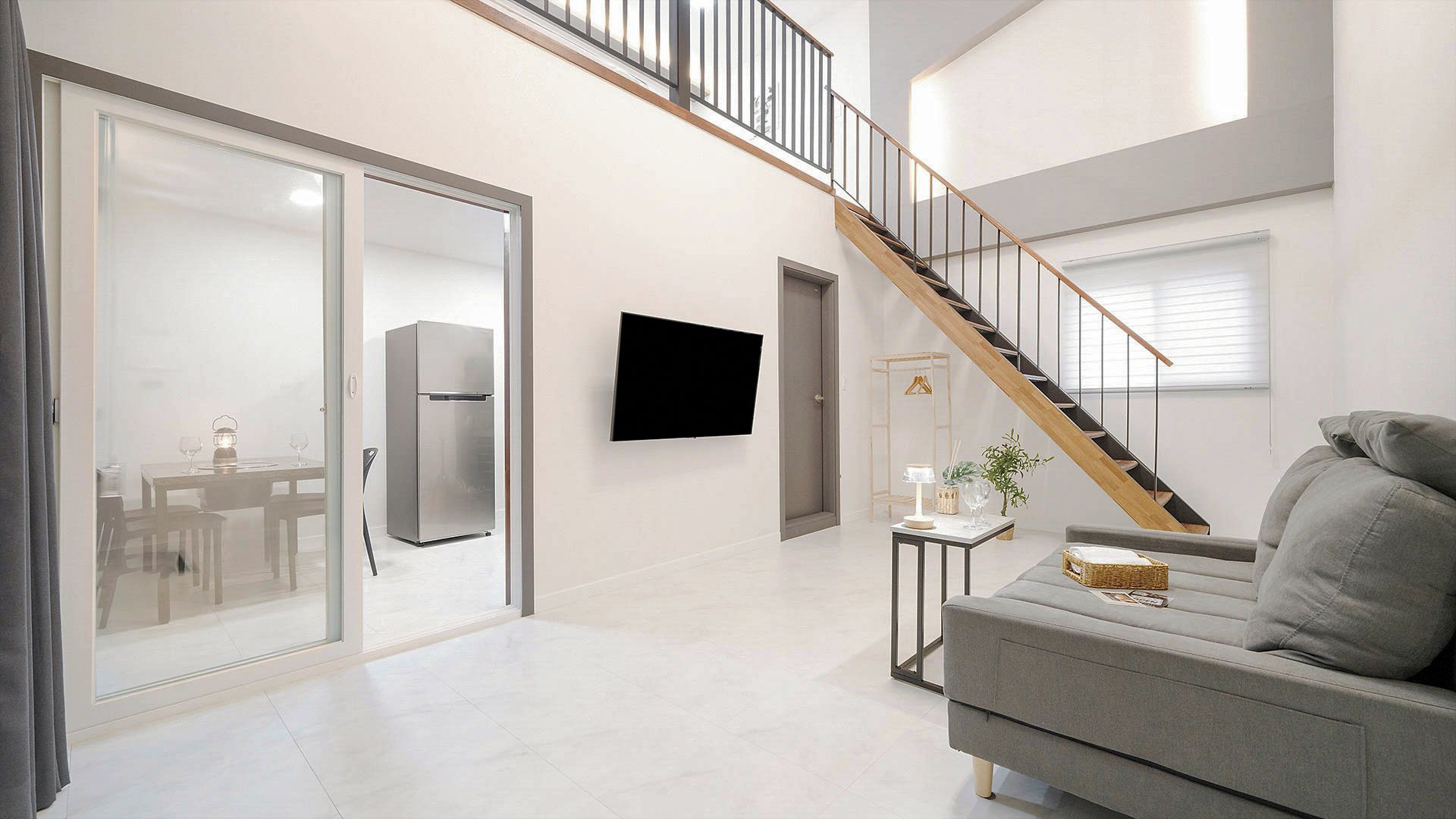 Una sala de estar moderna con un sofá gris, un televisor de pantalla plana, una escalera y un comedor con refrigerador.