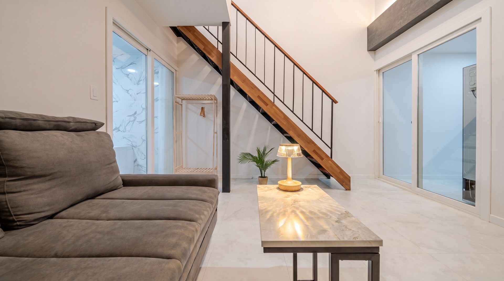 A modern living room with a sofa, a table with a lamp, and a staircase.