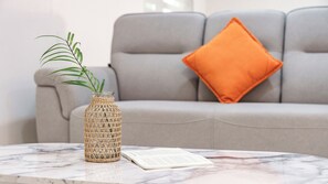 Un vase en osier avec une plante posé sur une table en marbre, un livre et un canapé gris avec un coussin orange.