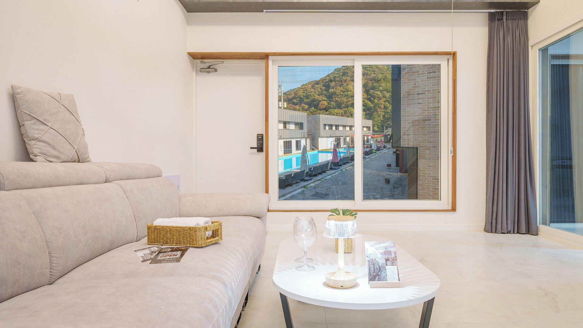 Una sala de estar moderna con un sofá, una mesa de centro con un florero de vidrio, una cesta de mimbre y un cuadro con una vista de un área de piscina.