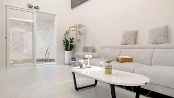 A modern living room with a white sofa, a glass door leading to a bathroom, and a coffee table with decorative items.