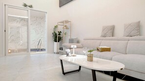 Ein modernes Wohnzimmer mit einem weißen Sofa, einer Glastür zu einem Badezimmer und einem Couchtisch mit Dekorationselementen.