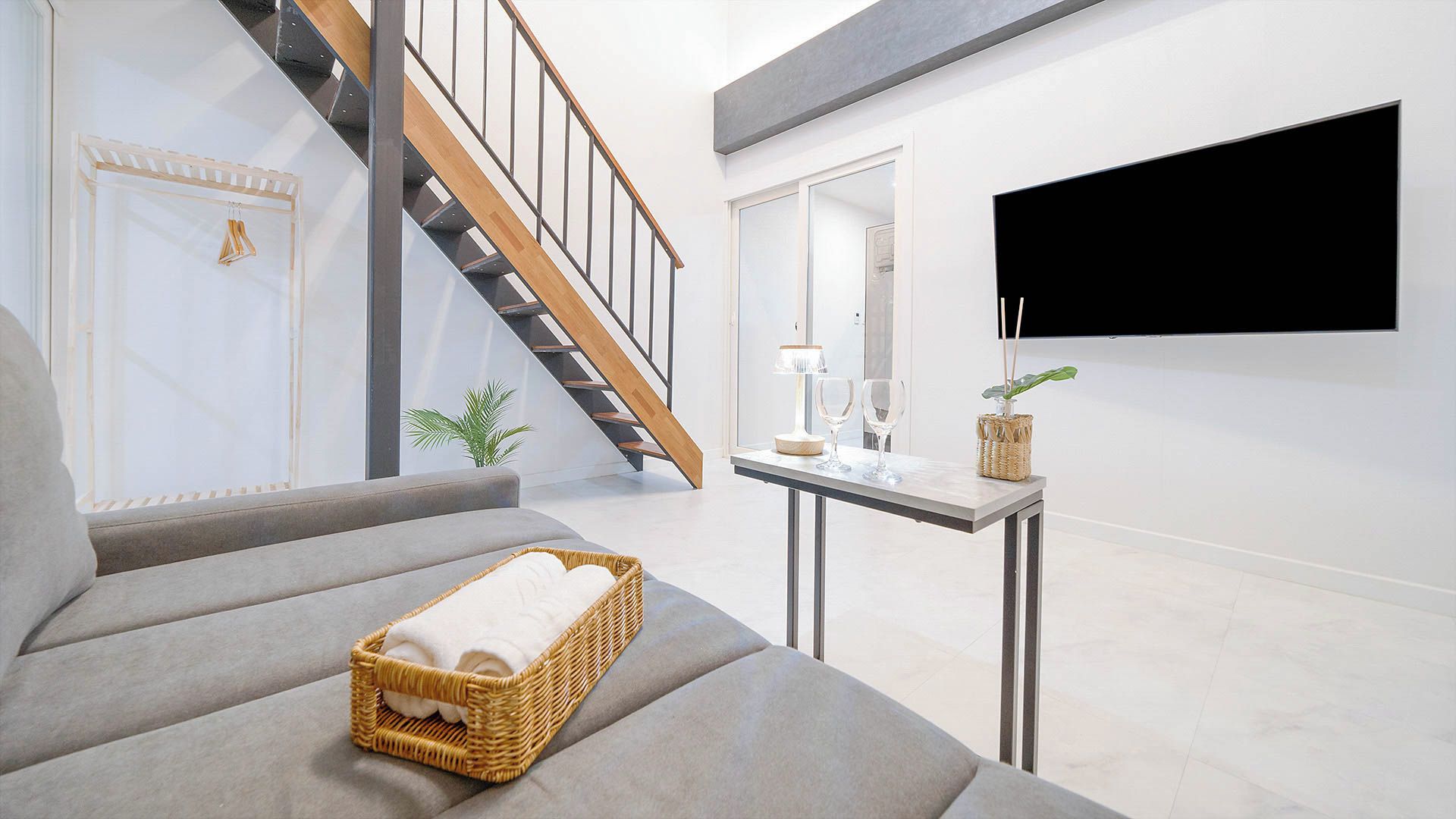 A modern living room with a grey sofa, a wicker basket with a towel, a small table with a lamp and glasses, and a TV mounted on the wall.