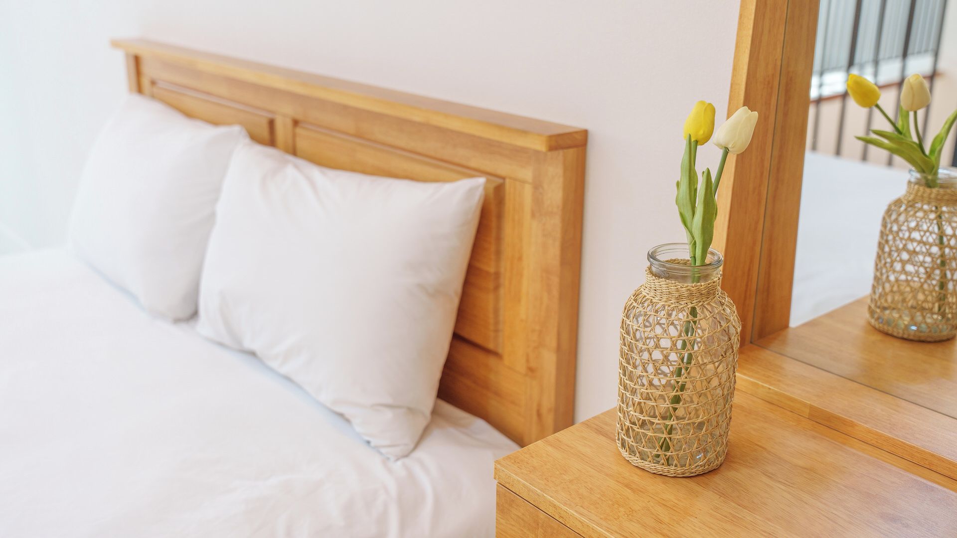 A bed with white bedding, a wooden headboard, and a nightstand with a woven glass vase containing flowers.