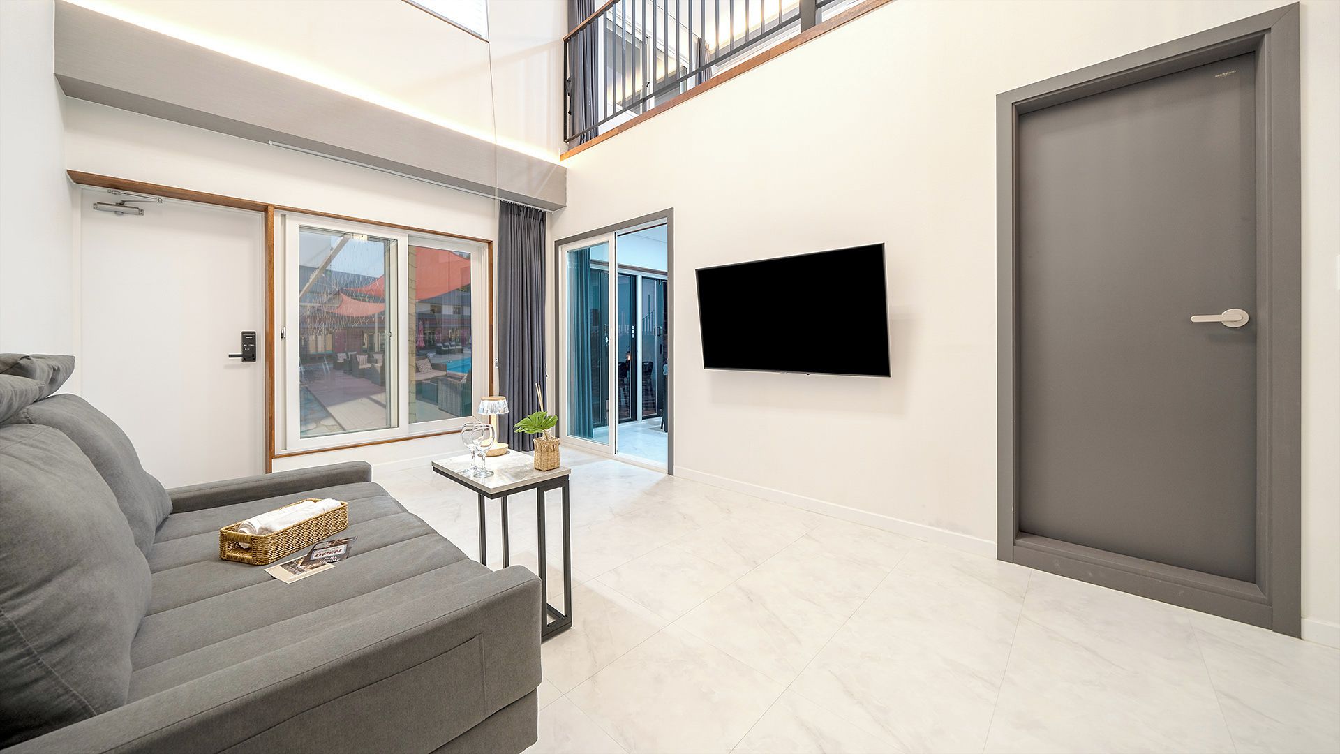 A modern living room with a grey sofa, a small table with a lamp, and a TV mounted on the wall.
