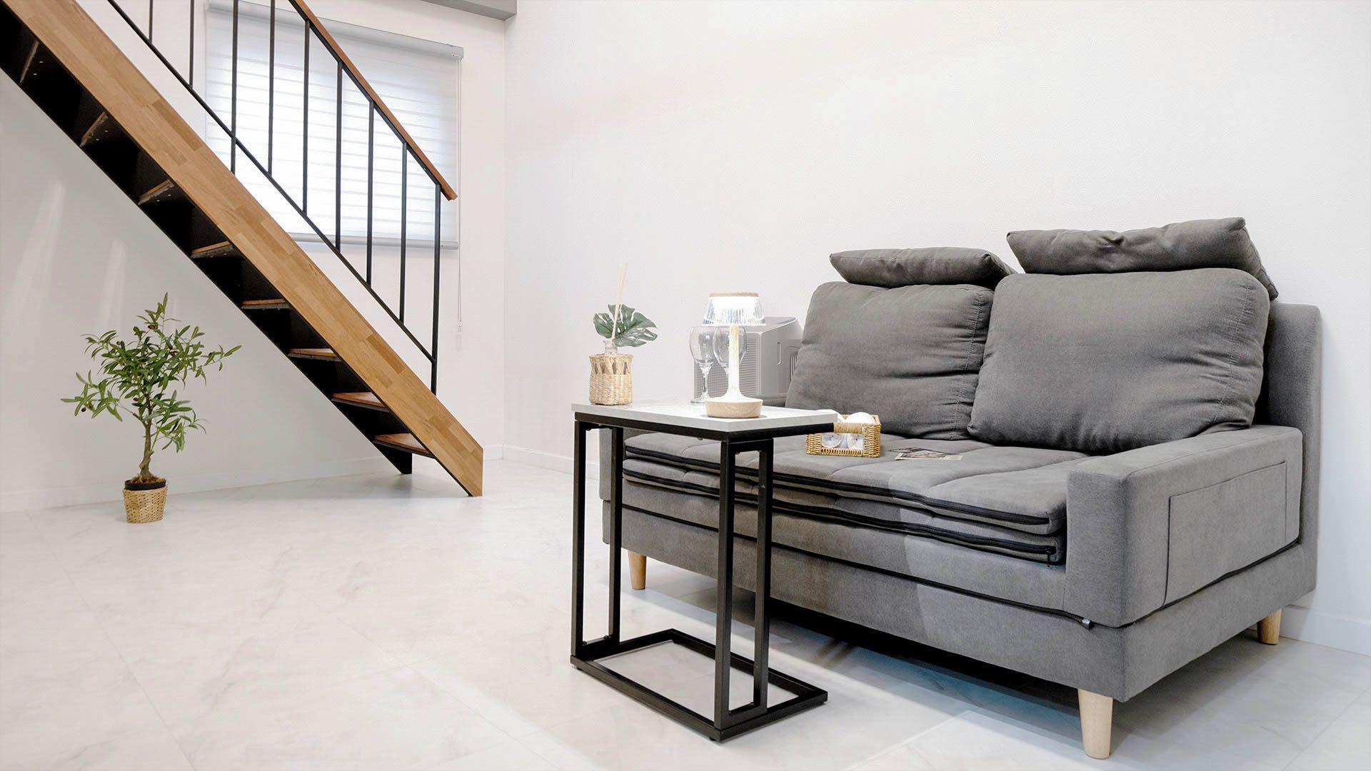 A modern living room with a grey sofa, a small table, a potted plant, and a wooden staircase.