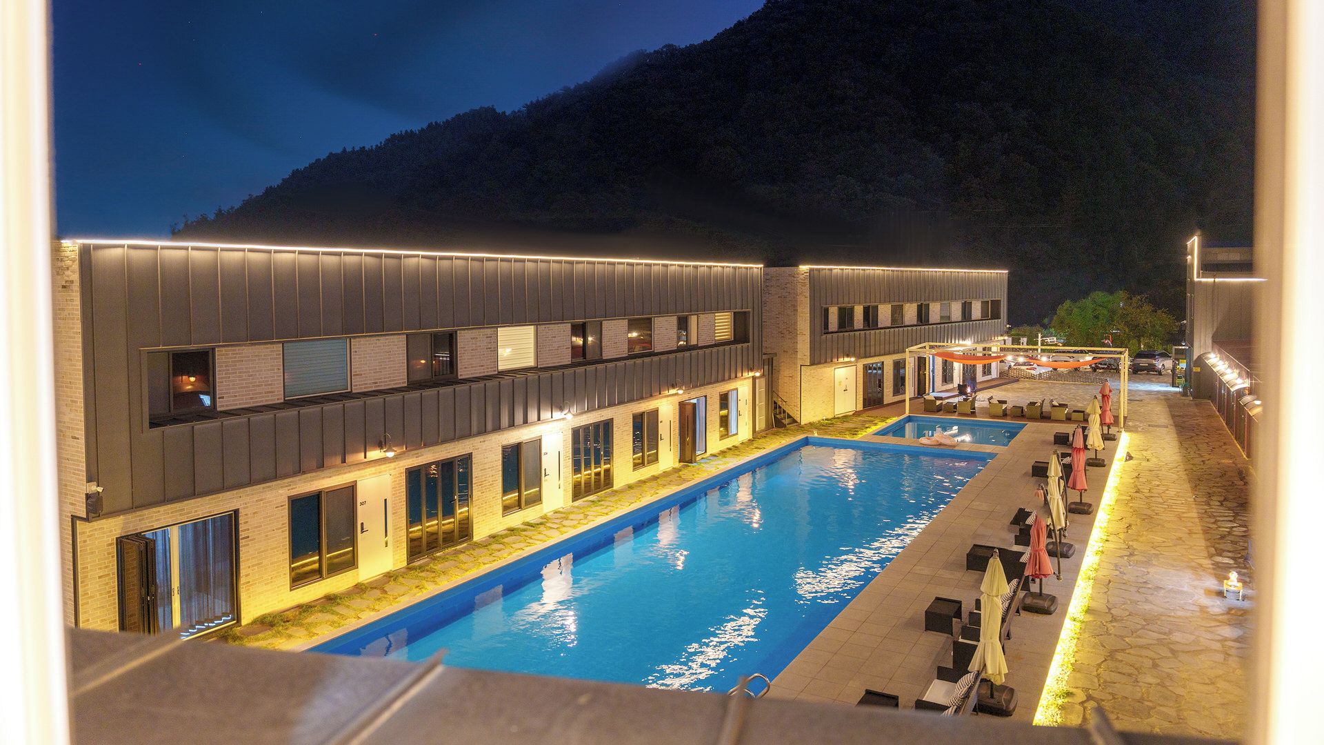 A modern hotel with a swimming pool, multiple balconies, and a mountain backdrop.