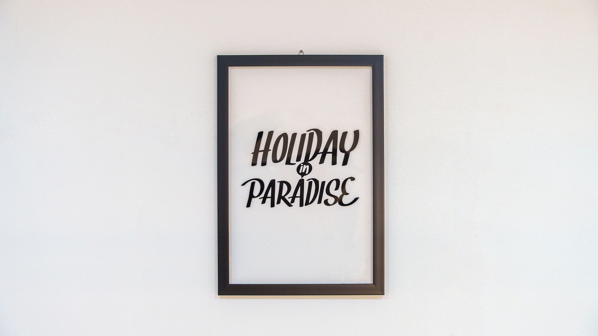 Un letrero en marco con el texto 'HOLIDAY in PARADISE' sobre una pared lisa.