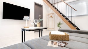 A modern living room with a grey sofa, a small table with a lamp and wine glasses, a wicker basket, and a staircase.