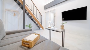 A modern living room with a grey sofa, a wicker basket with towels, a small table with a lamp and glasses, and a TV mounted on the wall.
