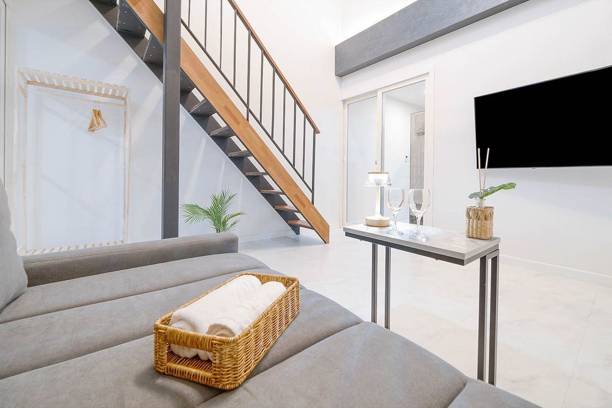 Un salon moderne avec un canapé gris, un panier en osier contenant des serviettes, une petite table avec une lampe et des verres, et un téléviseur fixé au mur.