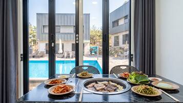 A dining table with various dishes, a grill, and a view of a swimming pool and buildings.