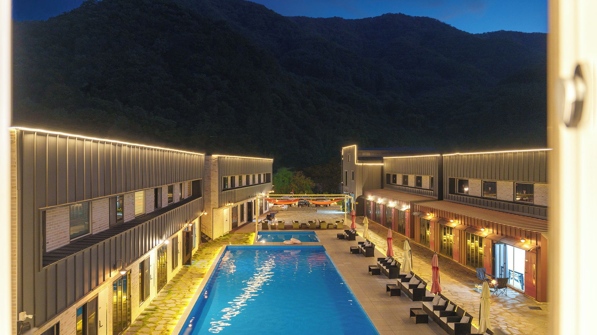 A poolside area with a pool, lounge chairs, and a view of mountains at night.
