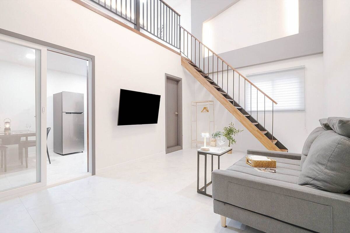 Un salon moderne avec un canapé gris, un téléviseur à écran plat, un escalier et un coin repas visible à travers une porte en verre.