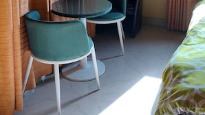 Une petite pièce avec une table ronde et deux chaises, une chaise noire, un appareil de climatisation et un lit recouvert d’une courtepointe à motifs.