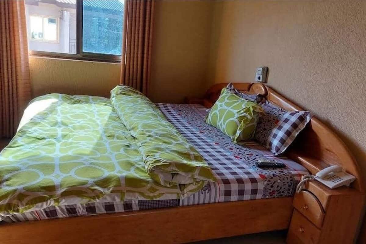 A single bed with a patterned bedspread and pillows, a bedside telephone, and a window with curtains.