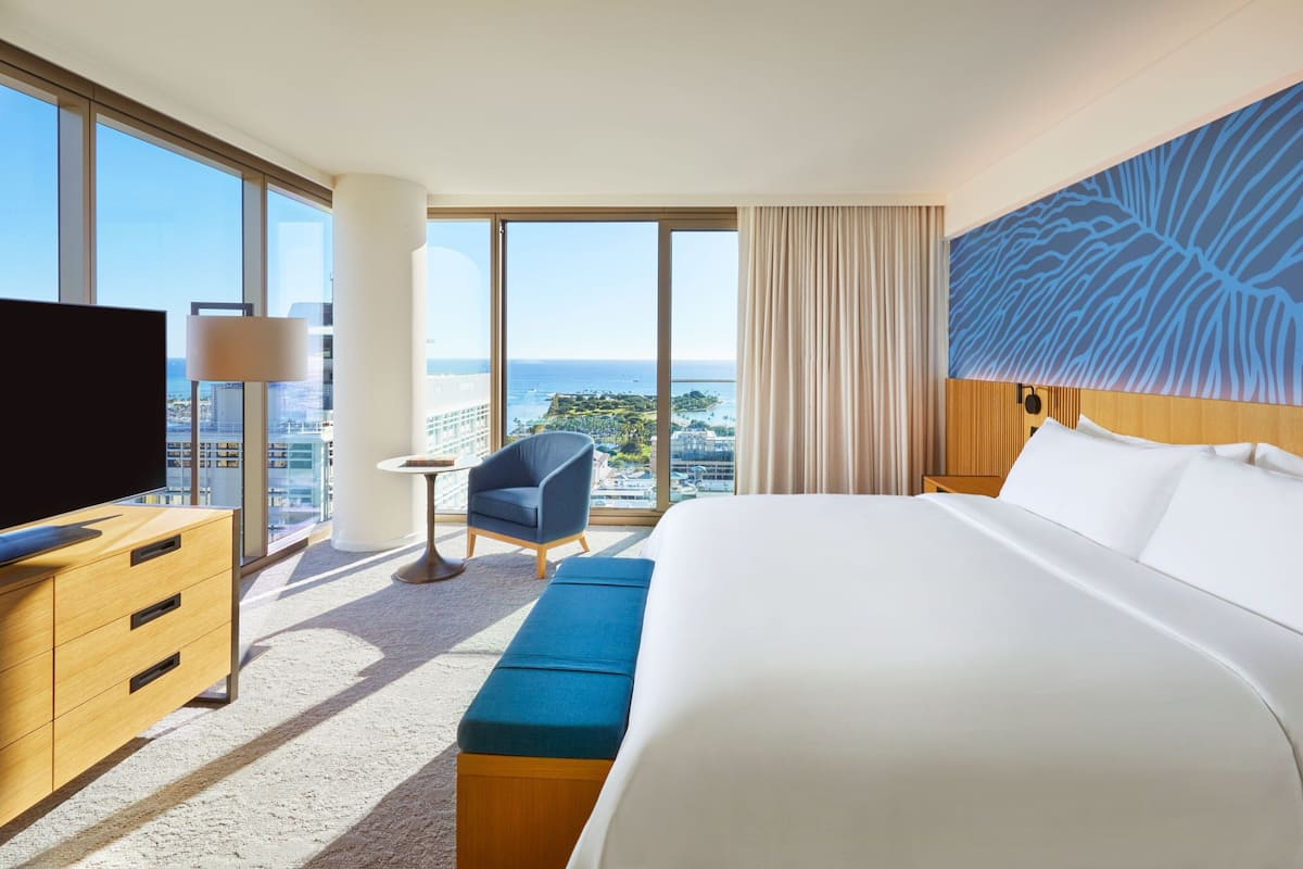 A hotel room with a large bed, a TV, a desk, a chair, and a view of the city and ocean.