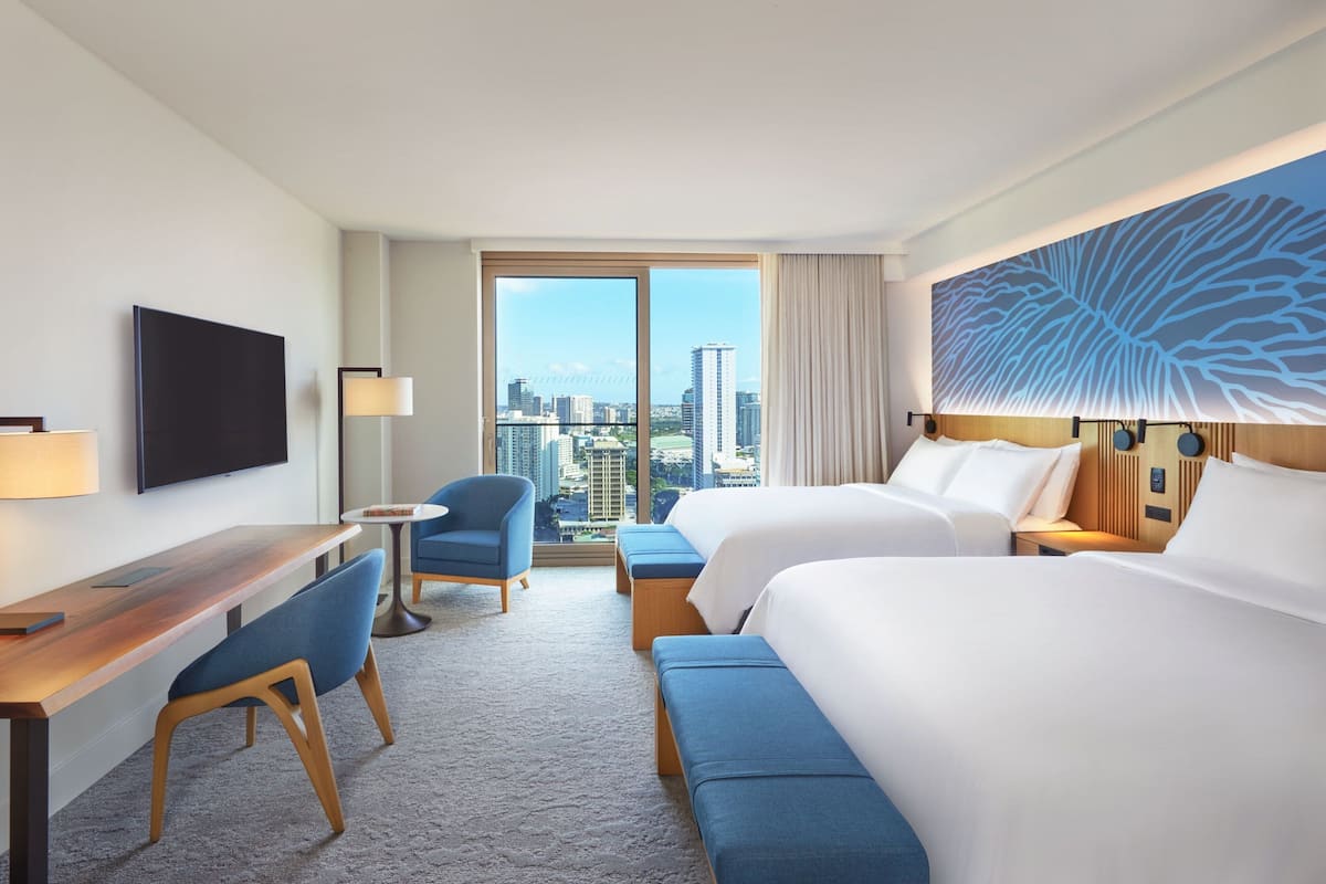 A hotel room with a large bed, a desk, a chair, and a TV. There is a window with a city view.