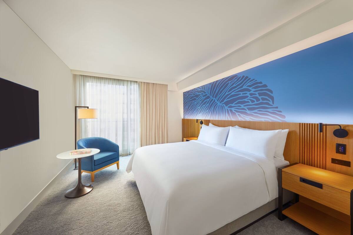 A hotel room with a large bed, a small round table, a blue chair, a nightstand, a lamp, and a large wall mural.