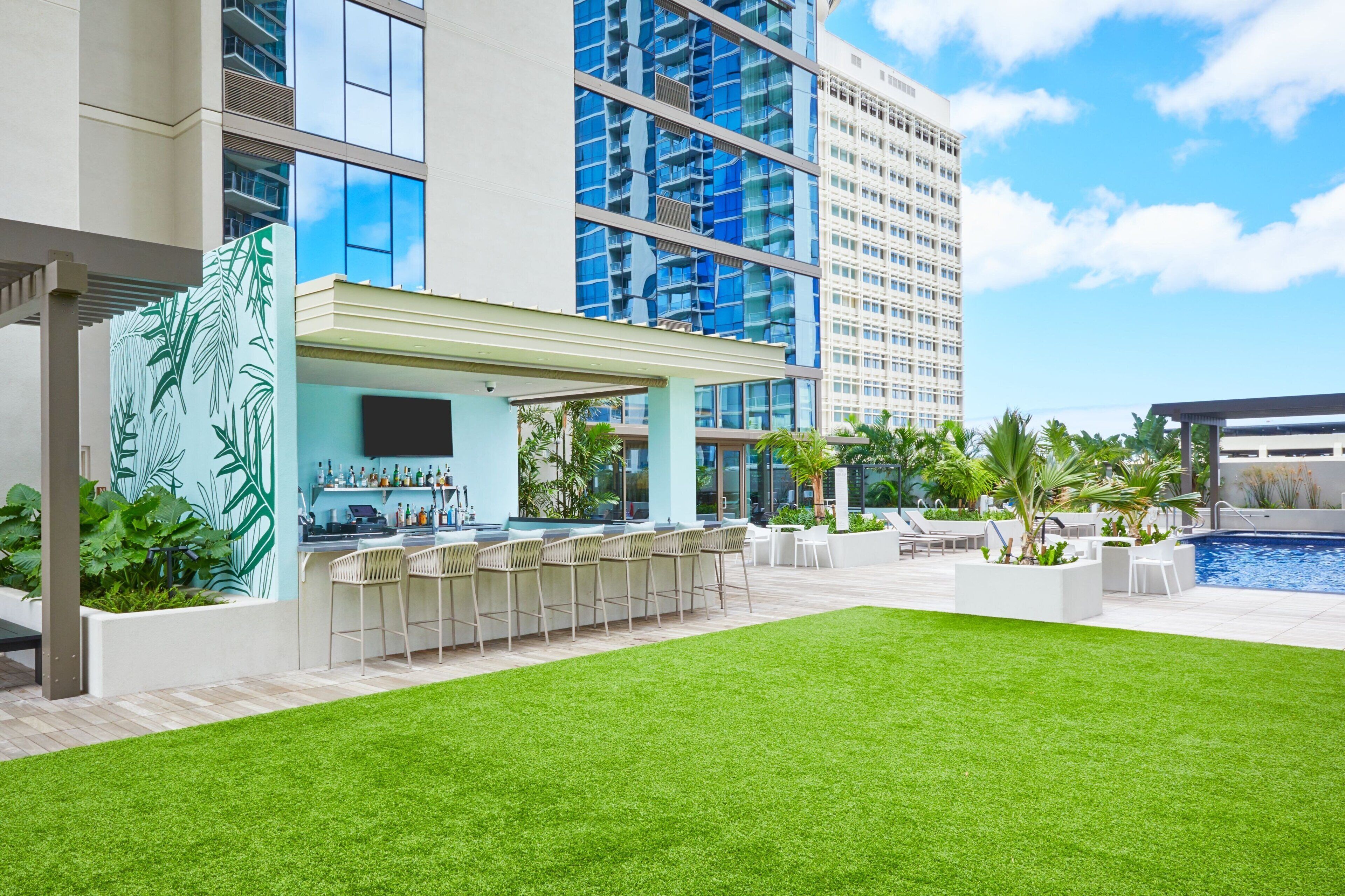 Un espace bar extérieur moderne, doté d’une piscine, entouré d’une végétation luxuriante et d’une pelouse bien entretenue.