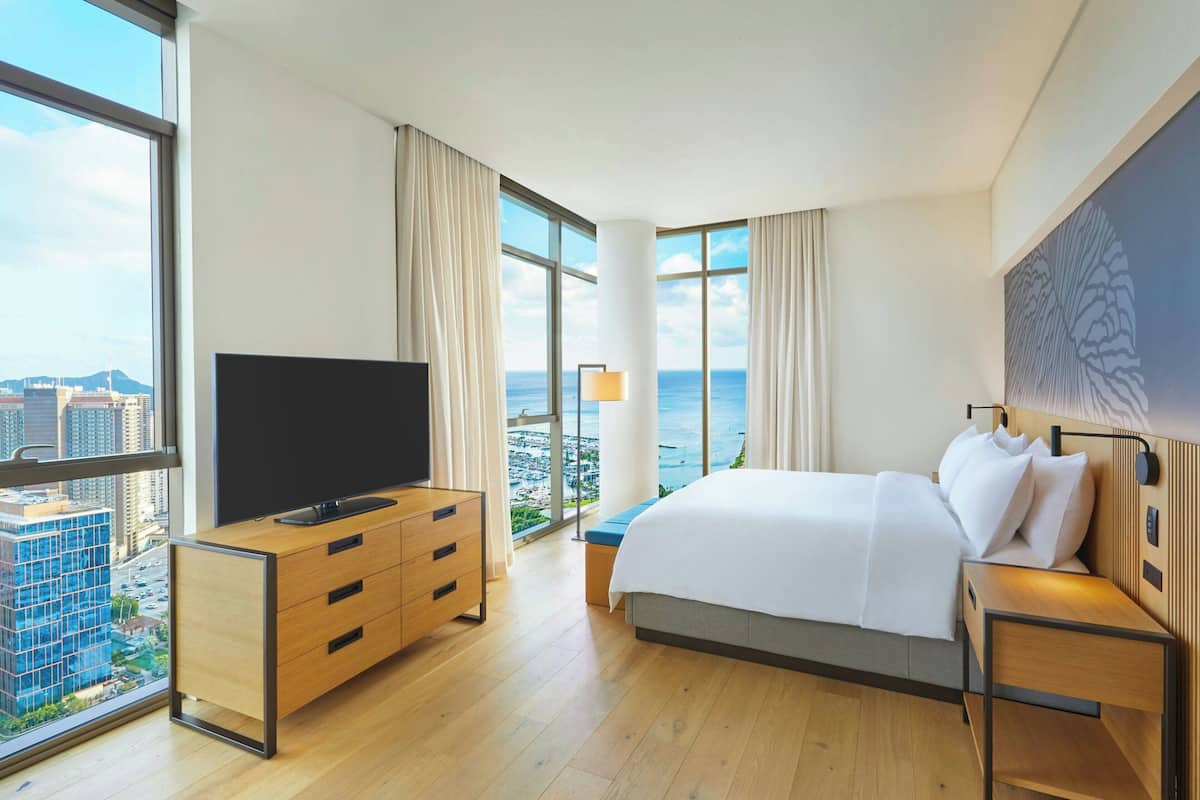 A modern hotel room with a large bed, a TV on a wooden dresser, and a city view through large windows.