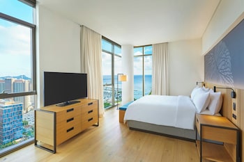 A hotel room with a large bed, a TV, a desk, a chair, and a view of the city and ocean. at Renaissance Honolulu Hotel & Spa