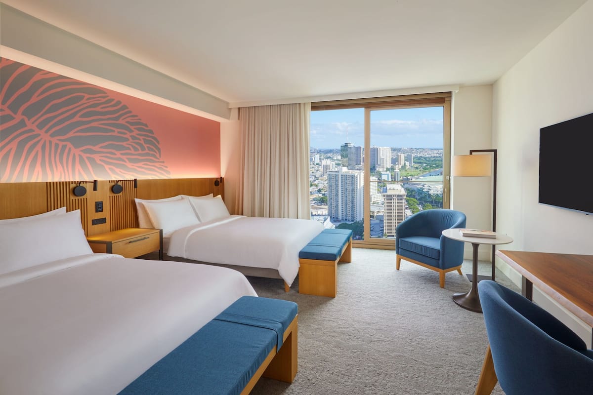 A hotel room with two beds, a desk, a chair, and a view of the cityscape.