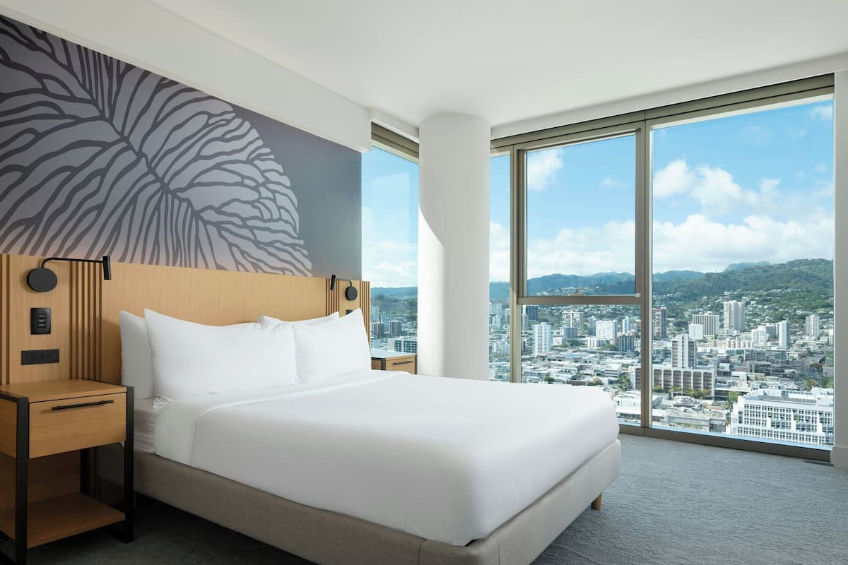A hotel room with a large bed, a nightstand, a wall-mounted lamp, and a city view through large windows.