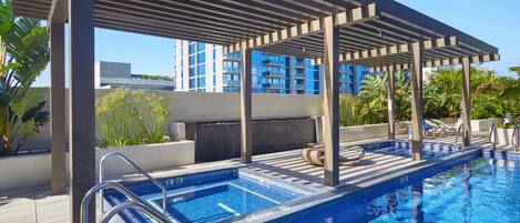 A rooftop swimming pool with a pergola, lounge chairs, and a view of high-rise buildings.