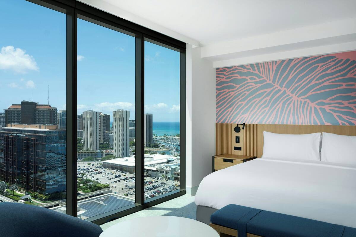 Quarto de hotel com uma janela ampla que oferece vista para a cidade, uma cama com cabeceira estampada e uma mesa pequena.
