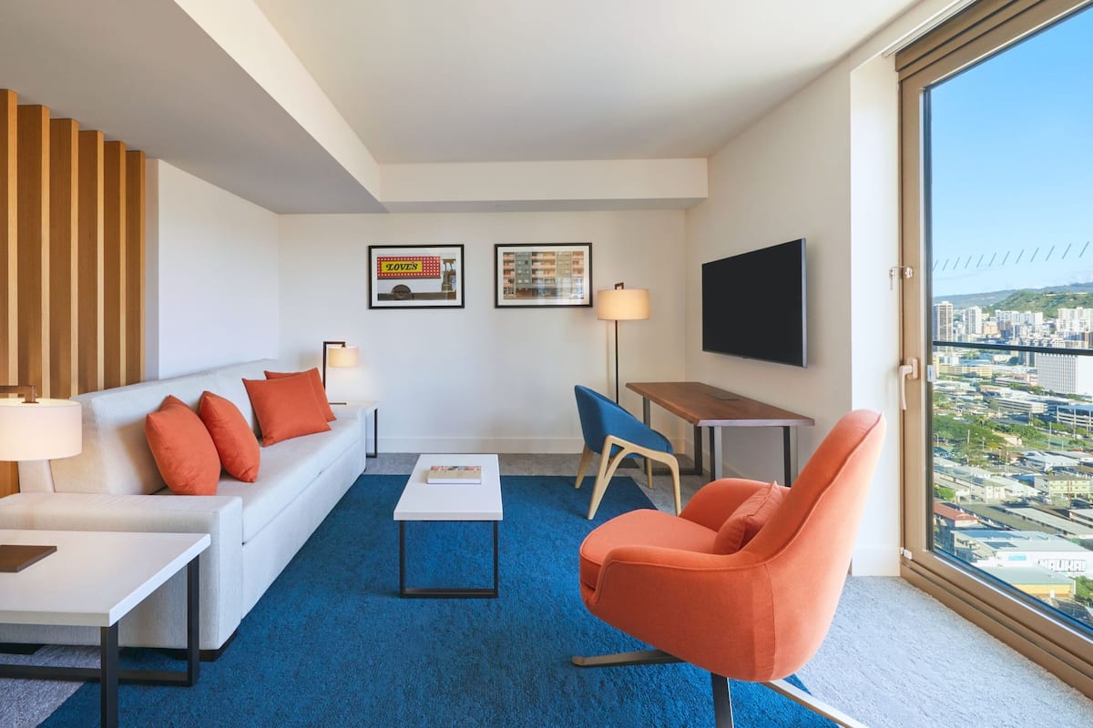 A modern hotel room with a sofa, armchair, coffee table, and a TV.