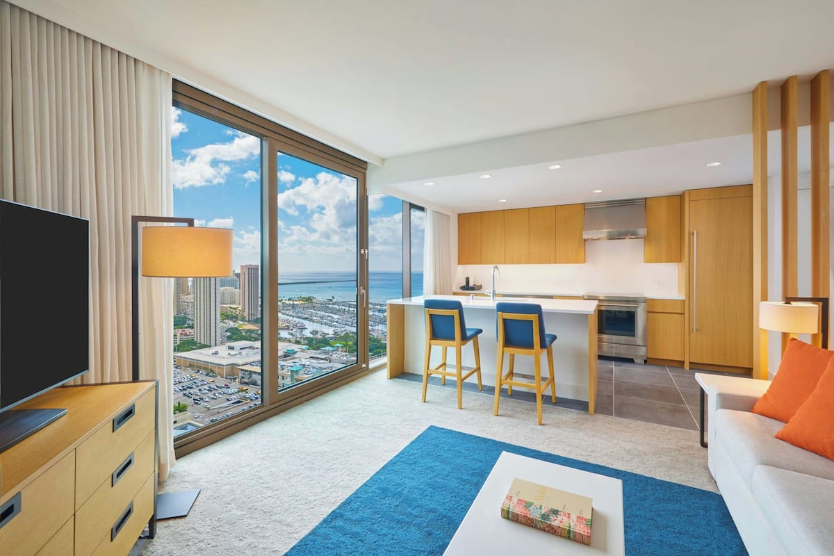 A modern hotel room with a large window offering a city view, a kitchenette, a flat-screen TV, a sofa, and a coffee table.