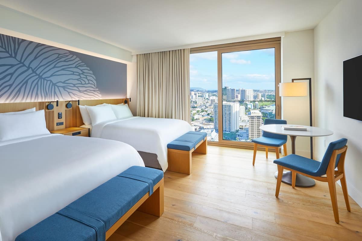 A hotel room with two beds, a dining area, and a large window offering a city view.